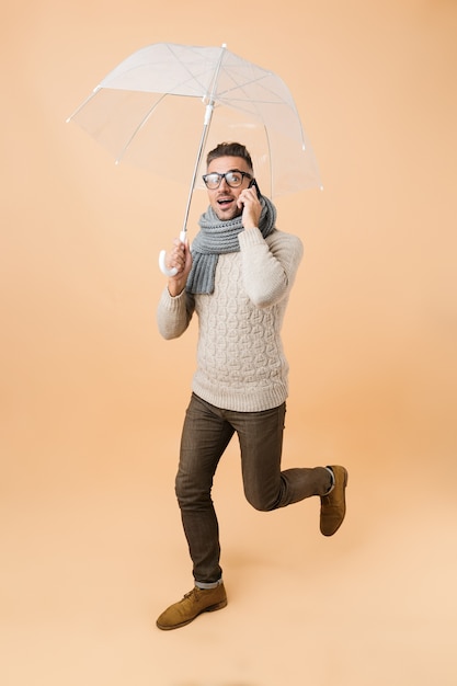 Retrato de cuerpo entero si un hombre guapo vestido con suéter y bufanda caminando bajo el paraguas aislado sobre la pared beige, hablando por teléfono móvil