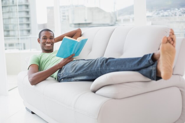 Retrato de cuerpo entero de sonriente hombre afro leyendo un libro en el sofá