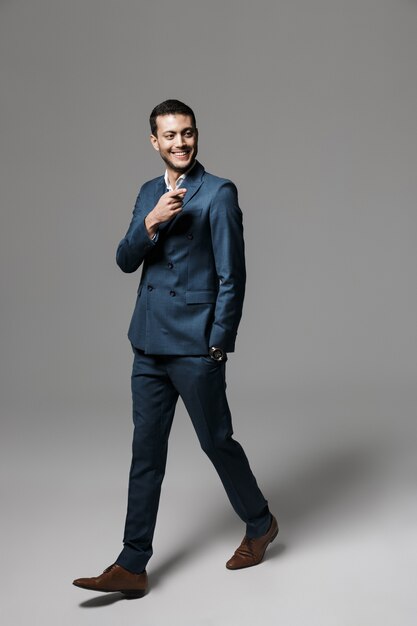 Retrato de cuerpo entero de un sonriente joven empresario vestido con traje caminando aislado sobre pared gris, apuntando hacia afuera