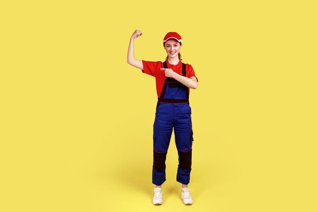 Retrato de cuerpo entero de una trabajadora optimista de pie con los brazos levantados mostrando su poder apuntando a sus bíceps con un mono y una gorra roja Toma de estudio interior aislada en fondo amarillo