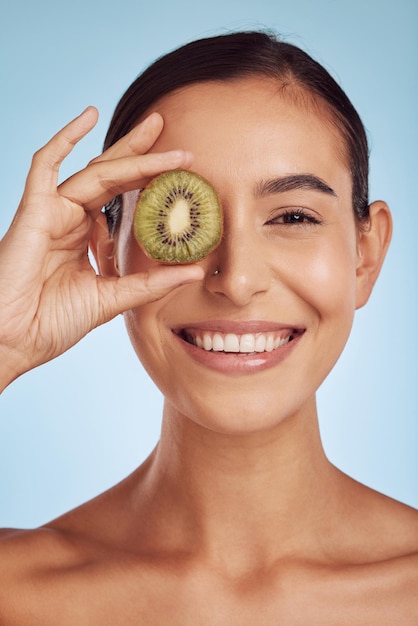 Retrato de cuidado de la piel kiwi y mujer con cosméticos de belleza para los ojos y salud de productos naturales y vitamina c Cara de persona joven feliz o modelo de frutas verdes y dermatología sobre un fondo azul de estudio