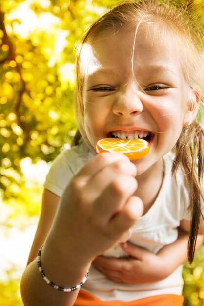 Retrato de Cute Little Girl de pie en el parque y sosteniendo en la mano Lollipop naranja.