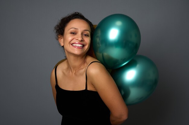 Retrato da altura da cintura de uma mulher bonita com um lindo sorriso segurando bolas de ar brilhantes atrás das costas e olhando para a câmera contra um fundo de parede cinza com espaço de cópia para o anúncio de Natal ou evento festivo