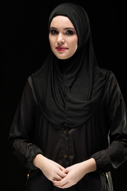 Retrato da bela jovem muçulmana positiva vestindo preto hijab como conceito de moda conservadora com a mão na mão, sorrindo em fundo preto