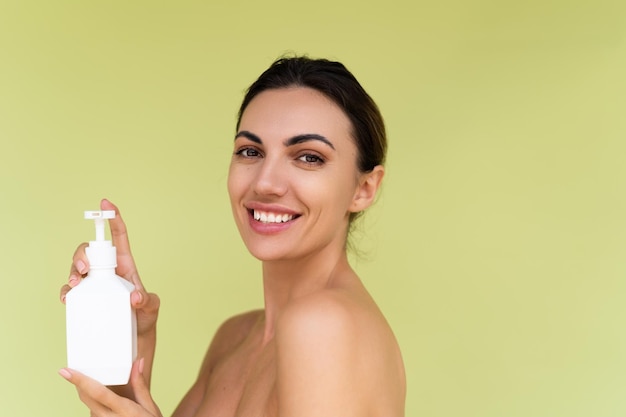 Retrato da beleza da jovem mulher em topless com ombros nus em fundo verde com pele perfeita e maquiagem natural, segurando o frasco de loção para o corpo xampu