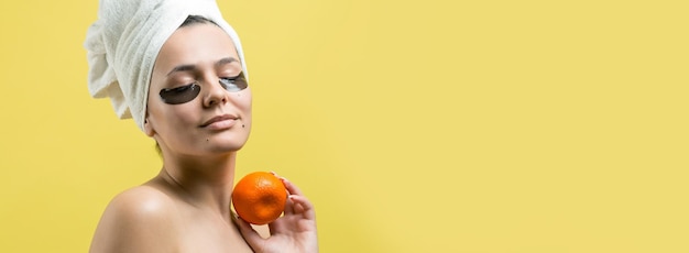 Retrato da beleza da mulher na toalha branca na cabeça com máscara nutritiva de ouro no rosto Limpeza de pele eco spa cosmético orgânico relax conceito Uma garota fica de costas segurando um mandarim laranja