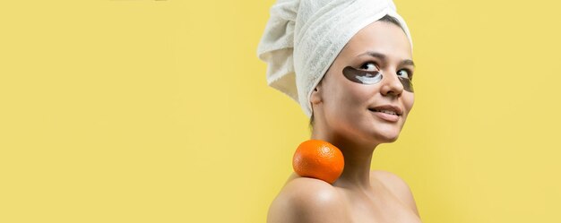 Retrato da beleza da mulher na toalha branca na cabeça com máscara nutritiva de ouro no rosto Limpeza de pele eco spa cosmético orgânico relax conceito Uma garota fica de costas segurando um tangerina laranja