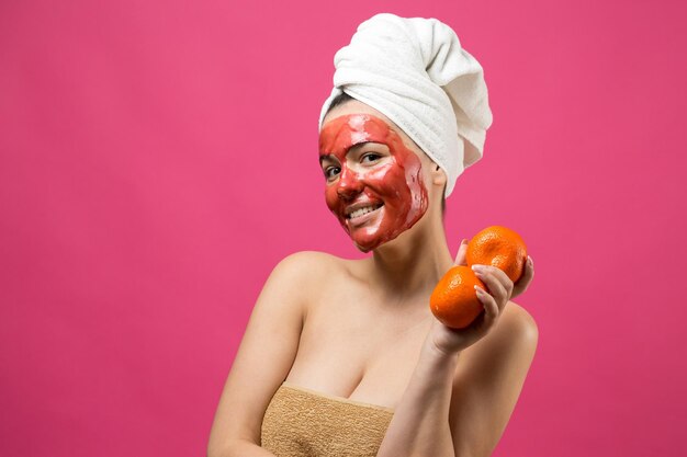 Retrato da beleza da mulher na toalha branca na cabeça com máscara nutritiva vermelha no rosto Skincare limpeza eco spa cosmético orgânico relax conceito Uma garota fica de costas segurando um mandarim laranja