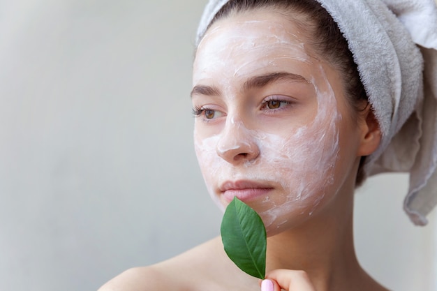 Retrato da beleza da mulher na toalha na cabeça com máscara nutritiva branca ou creme no rosto e folha verde na mão, fundo branco isolado. Conceito de spa cosmético ecológico de limpeza de cuidados da pele