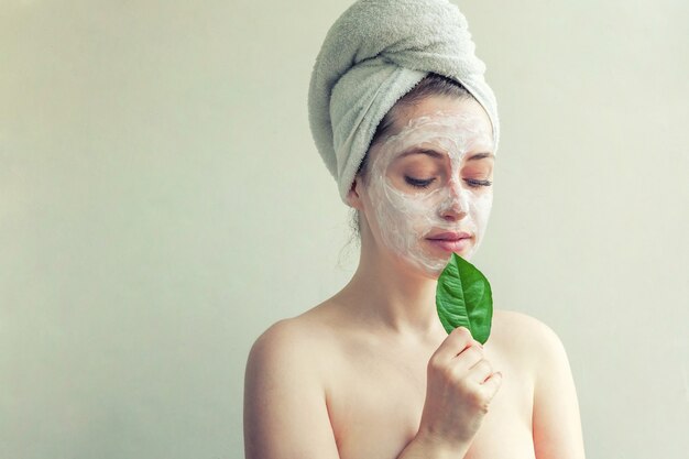 Retrato da beleza da mulher na toalha na cabeça com máscara nutritiva branca ou creme no rosto e folha verde na mão, fundo branco isolado. conceito de spa cosmético ecológico de limpeza de cuidados da pele