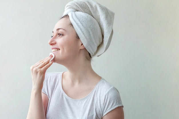 Retrato da beleza da mulher sorridente em uma toalha na cabeça com pele macia e saudável, removendo a maquiagem com almofada de algodão, isolada no fundo branco.