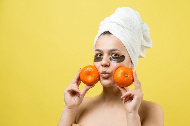 Retrato da beleza de mulher em uma toalha branca na cabeça com uma máscara nutritiva de ouro no rosto. Skincare cleansing eco orgânico cosmético spa relaxa conceito. Uma garota está de costas segurando uma tangerina laranja.