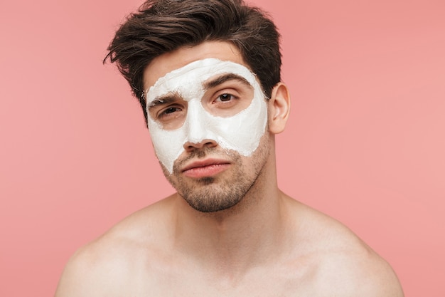 Retrato da beleza de um homem bonito, moreno sem camisa, isolado sobre uma parede rosa, usando máscara facial de cosmética