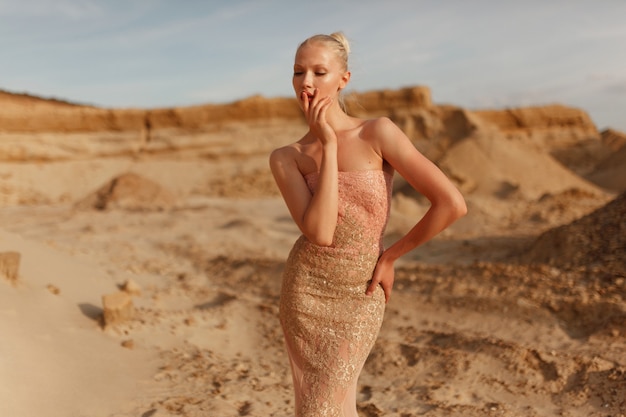 Retrato da beleza de uma jovem elegante vestida com um lindo vestido dourado dourado, posando no deserto ao pôr do sol.