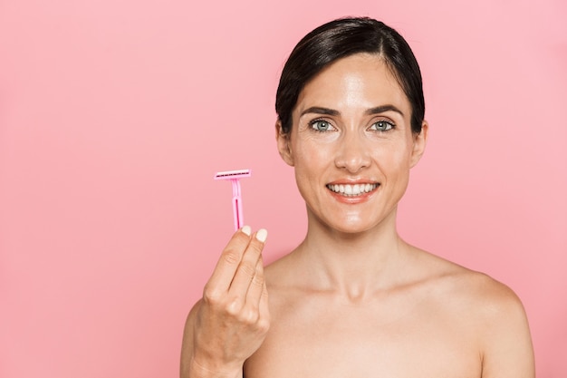 Retrato da beleza de uma jovem feliz e atraente em topless em pé isolado sobre uma parede rosa, segurando uma navalha