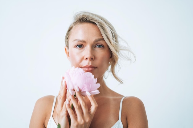 Retrato da beleza de uma jovem sorridente de cabelos loiros com peônia rosa na mão, isolada no fundo branco