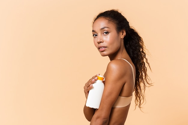 Retrato da beleza de uma jovem vestindo lingerie branca segurando um frasco com cosméticos, em pé, isolado sobre uma parede bege