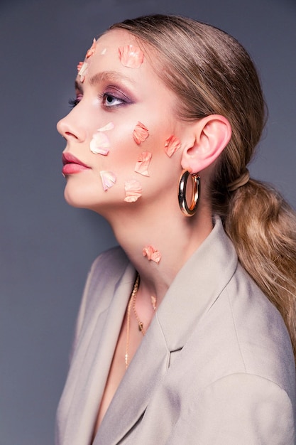 Retrato da beleza de uma menina mulher bonita em uma jaqueta posando com pétalas de flores no rosto em um fundo cinza feminilidade e beleza