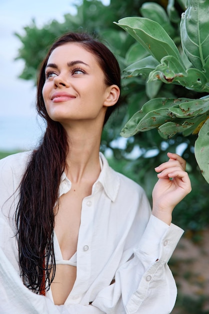 Foto retrato da beleza de uma mulher com cabelo molhado em uma camisa branca perto das folhas de uma árvore tropical beleza e saúde pele bronzeada o conceito de cuidados com a pele facial e saúde do cabelo nas férias