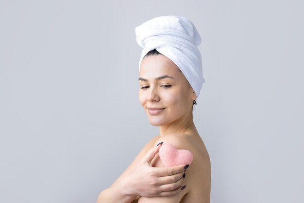 Retrato da beleza de uma mulher em uma toalha branca na cabeça com uma esponja para um corpo em vista de um coração rosa