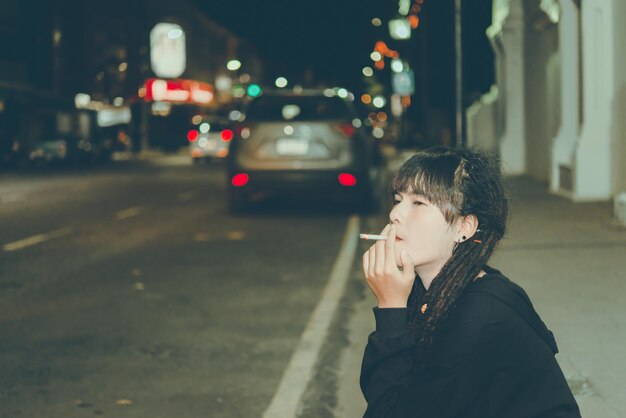 Retrato da cena noturna de fumar dreadlock de cabelo de mulher asiática na cidadeTailândia as pessoas posam para tirar um conceito de mulher triste solitária