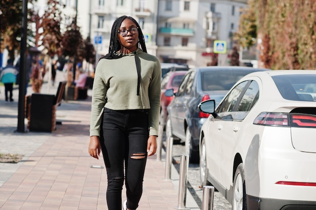Retrato da cidade de positivo jovem fêmea de pele escura, vestindo casaco com capuz verde e óculos andando no estacionamento.