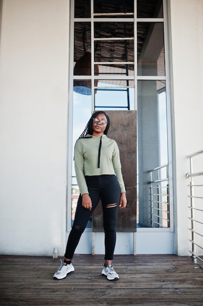 Retrato da cidade de uma jovem mulher de pele escura positiva vestindo capuz verde e óculos em pé em balkony