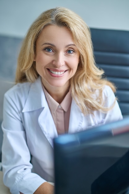 Retrato da cintura de uma encantadora médica loira de meia-idade com um computador olhando para a frente dela