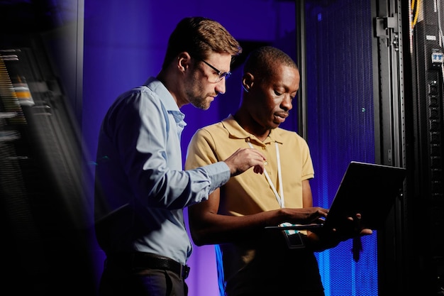 Retrato da cintura para cima de dois engenheiros de rede usando computador na sala escura do servidor iluminado por cópia de luz neon