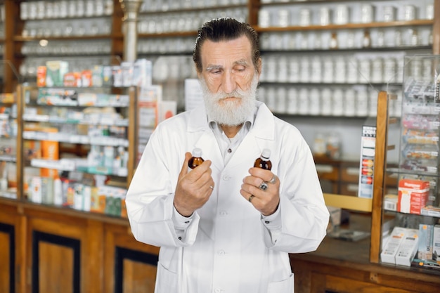 Retrato da cintura para cima de um belo farmacêutico idoso barbudo concentrado no trabalho na antiga farmácia comparando dois medicamentos segurando frascos nas mãos