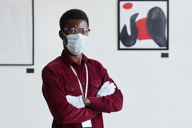 Retrato da cintura para cima de um homem afro-americano usando máscara, enquanto em pé com os braços cruzados contra pinturas gráficas modernas na galeria de arte,