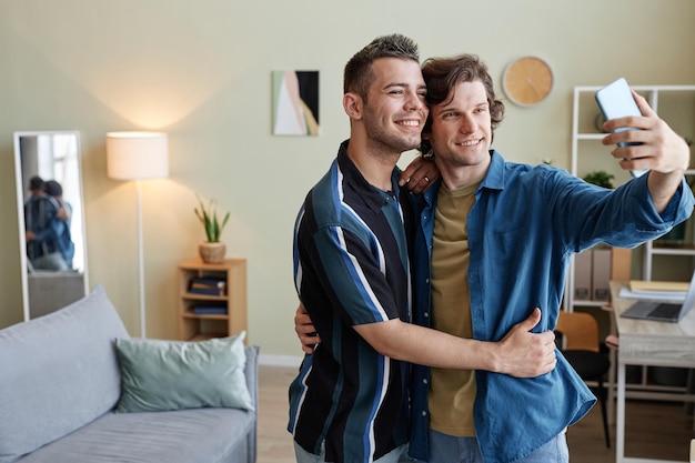 Retrato da cintura para cima de um jovem casal gay tirando fotos de selfie em uma nova casa juntos copiam o espaço