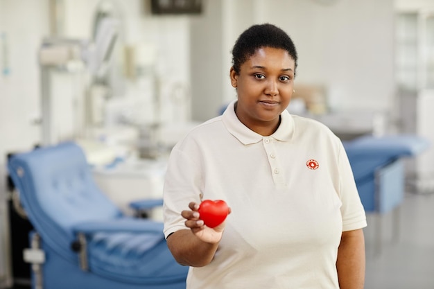 Retrato da cintura para cima de uma jovem negra segurando coração vermelho no centro de doação de sangue e olhando para a câmera