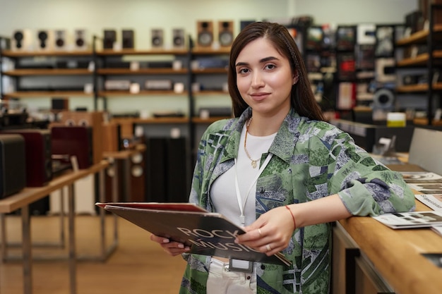 Retrato da cintura para cima de uma jovem trabalhando em uma loja de música e segurando um disco sorrindo para o espaço da cópia da câmera