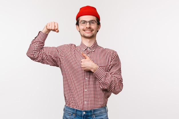 Retrato da cintura para cima do homem bonito feliz sorridente satisfeito com barba de óculos e gorro vermelho, flexione os bíceps e mostre o polegar para cima em aprovação ou algo assim, sorrindo orgulhosamente se gabando com sua força