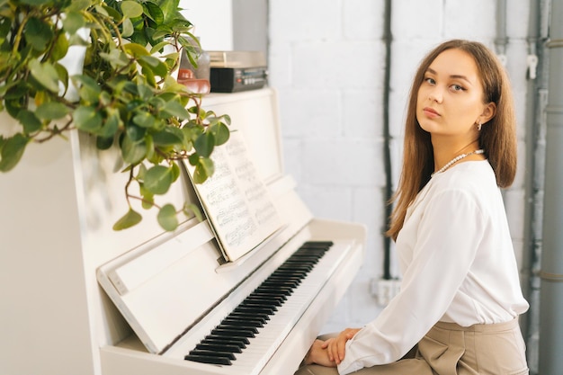 Retrato da elegância jovem pianista olhando para a câmera sentada no piano branco clássico em sala de aula Artista talentoso músico feminino se preparando para aulas em estúdio em casa