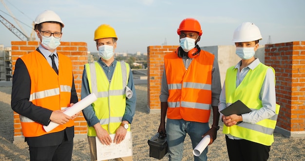 Retrato da equipe de construtores de homens e mulheres de raças mistas em capacetes e máscaras médicas em pé no topo do edifício com a construção de rascunhos de planos. Coronavírus. Engenheiros e arquitetos em construção.