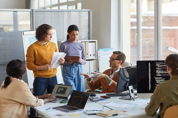 Retrato da equipe diversificada de desenvolvimento de software colaborando no projeto em um escritório moderno, foco no engenheiro líder instruindo colegas, copie o espaço