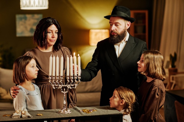 Retrato da família judaica ortodoxa acendendo a vela menorah durante a celebração do hanukkah em casa