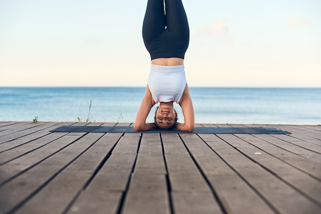 Retrato da ioga praticando da mulher bonita do ajuste contra o mar azul que está na pose do headstand.