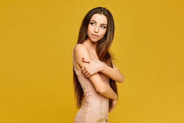 Retrato da jovem modelo mulher com corpo esguio e cabelo comprido, usando um vestido bege isolado no fundo amarelo com espaço de cópia