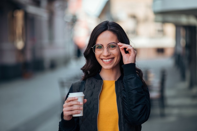 Retrato da jovem mulher de sorriso que guarda o copo de papel, cena urbana, vida urbana.