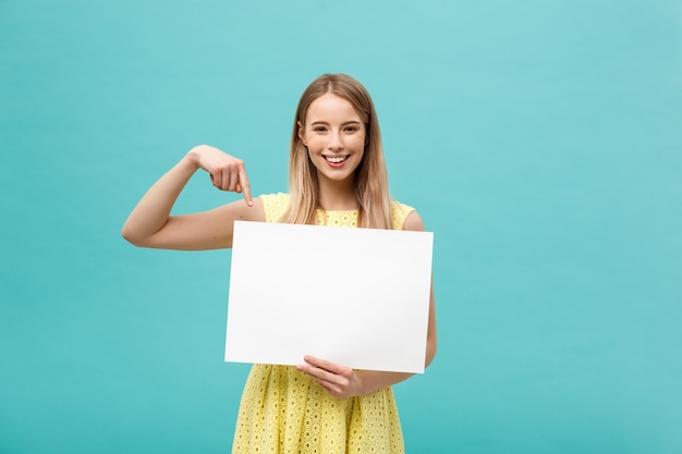 Retrato da jovem mulher no vestido amarelo que aponta o dedo na placa em branco branca lateral.