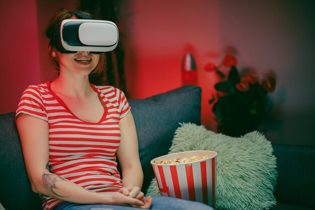 Retrato da jovem mulher sentada no sofá e com o fone de ouvido VR, assistindo algo enquanto come pipoca e sorrindo