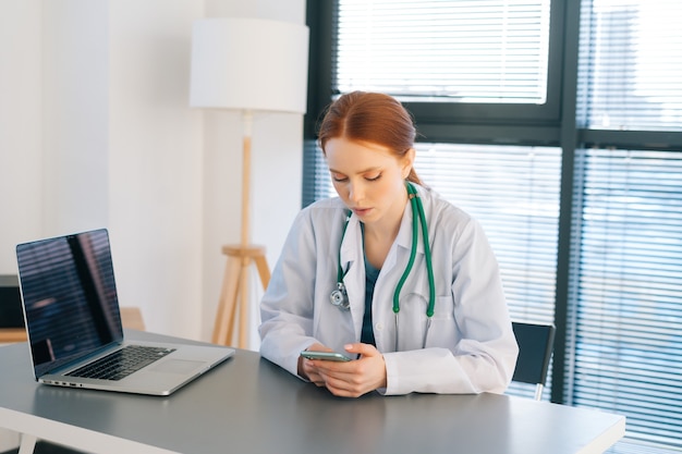 Retrato da médica ruiva atraente no jaleco branco, dando rápido diagnóstico remoto aos pacientes usando telefone celular, sentado na mesa com o laptop perto da janela em dia ensolarado.