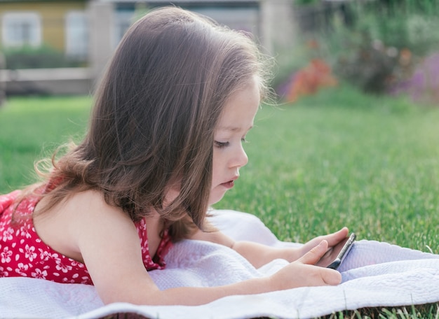 Retrato da menina 3-4 em vermelho deitado no cobertor na grama verde e olhando para o telefone móvel. crianças usando gadgets
