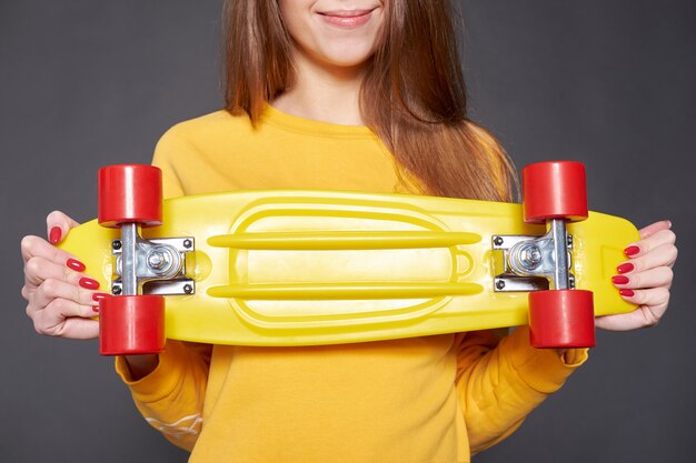 Retrato da menina atrativa nova positiva que veste a blusa amarela e a calças de ganga que guardam o skate amarelo.