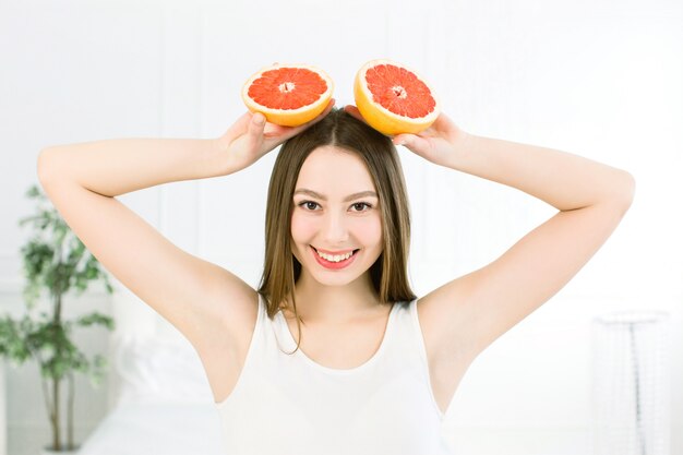 Retrato da menina consideravelmente positiva atrativa alegre que tem duas partes de toranja em suas mãos aéreas no fundo claro. Conceito de comida saudável. Vitaminas e minerais.