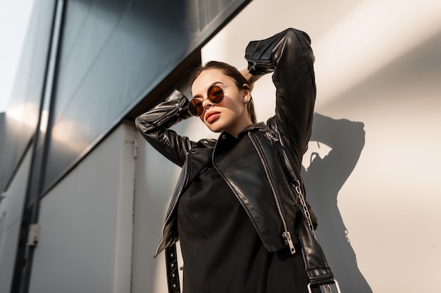 retrato da moda da bela modelo feminina com óculos de sol em elegantes roupas pretas, com jaqueta de couro na cidade na luz do sol. Garota fashion
