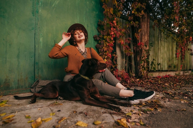 Retrato da moda da modelo sorridente com cabelo curto encaracolado e em roupas vintage senta-se com cachorro no outono no chão perto de chalé posa para a câmera com cara feliz modelo elegante na aldeia com um cachorro